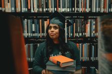 A graduate in a cap and gown holding books in a library