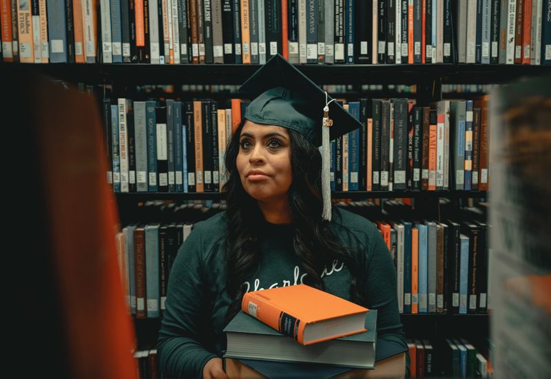 A graduate in a cap and gown holding books in a library