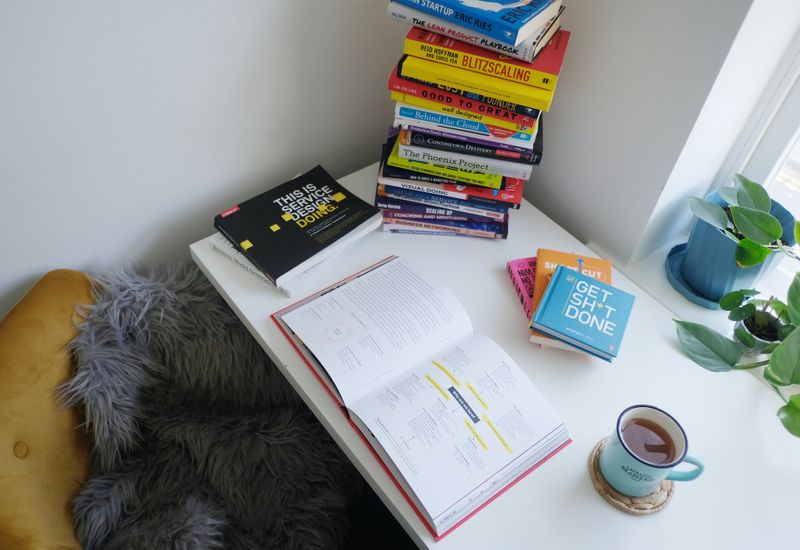Desk with a large pile of books, an open notebook, and a cup of coffee