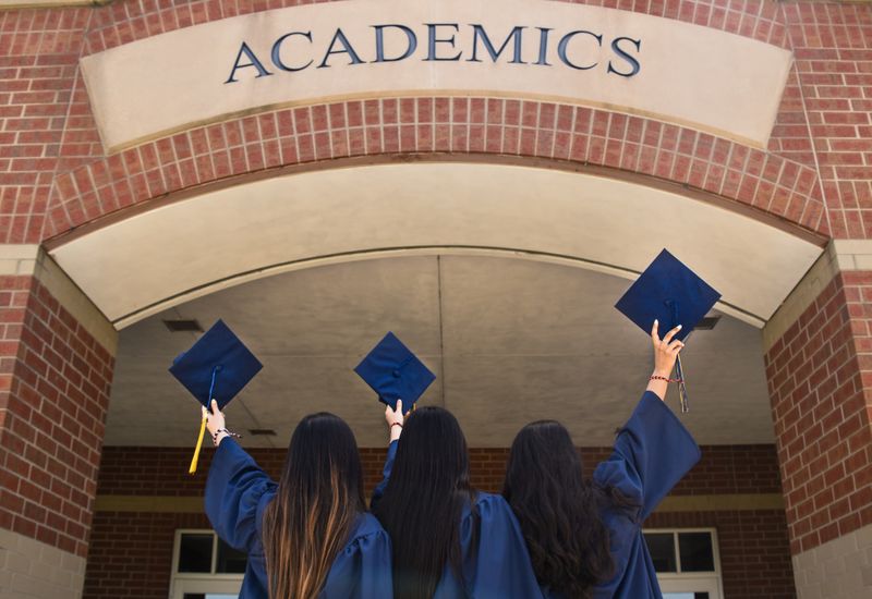 Three students holding their mortarboards up in front of a sign on a red brick building that says "Academics"