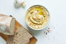 A bowl of hummus complete with olive oil, paprika and whole chickpeas as a garnish, with some sliced bread and a garlic bulb