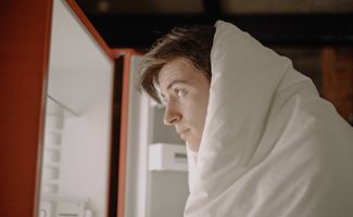 Young man wearing a duvet looking into an empty fridge