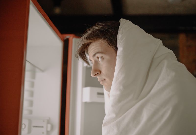 Young man wearing a duvet looking into an empty fridge