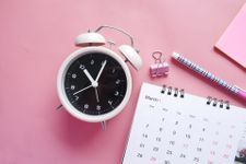 Traditional alarm clock, calendar, bulldog clip, pencil, and post-it on a pink desk